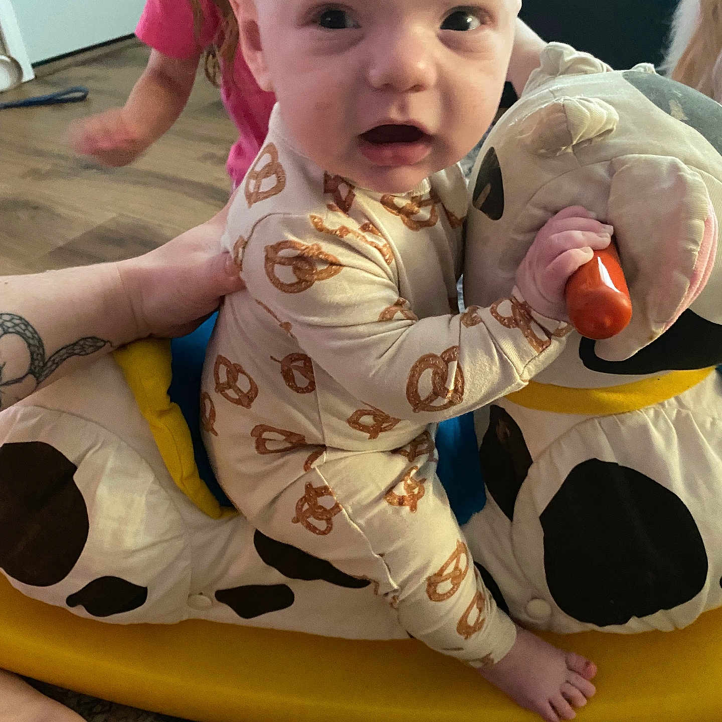 Wesley is registered to the contest to win money with this photo: baby, child, pretzel_pajamas, plush_toy, cow, yellow_rocker, indoor, floor, hand, person, foot, carpet, wood_floor, child_in_background, toy, clothing, supporting_hand, expression, face, sitting
