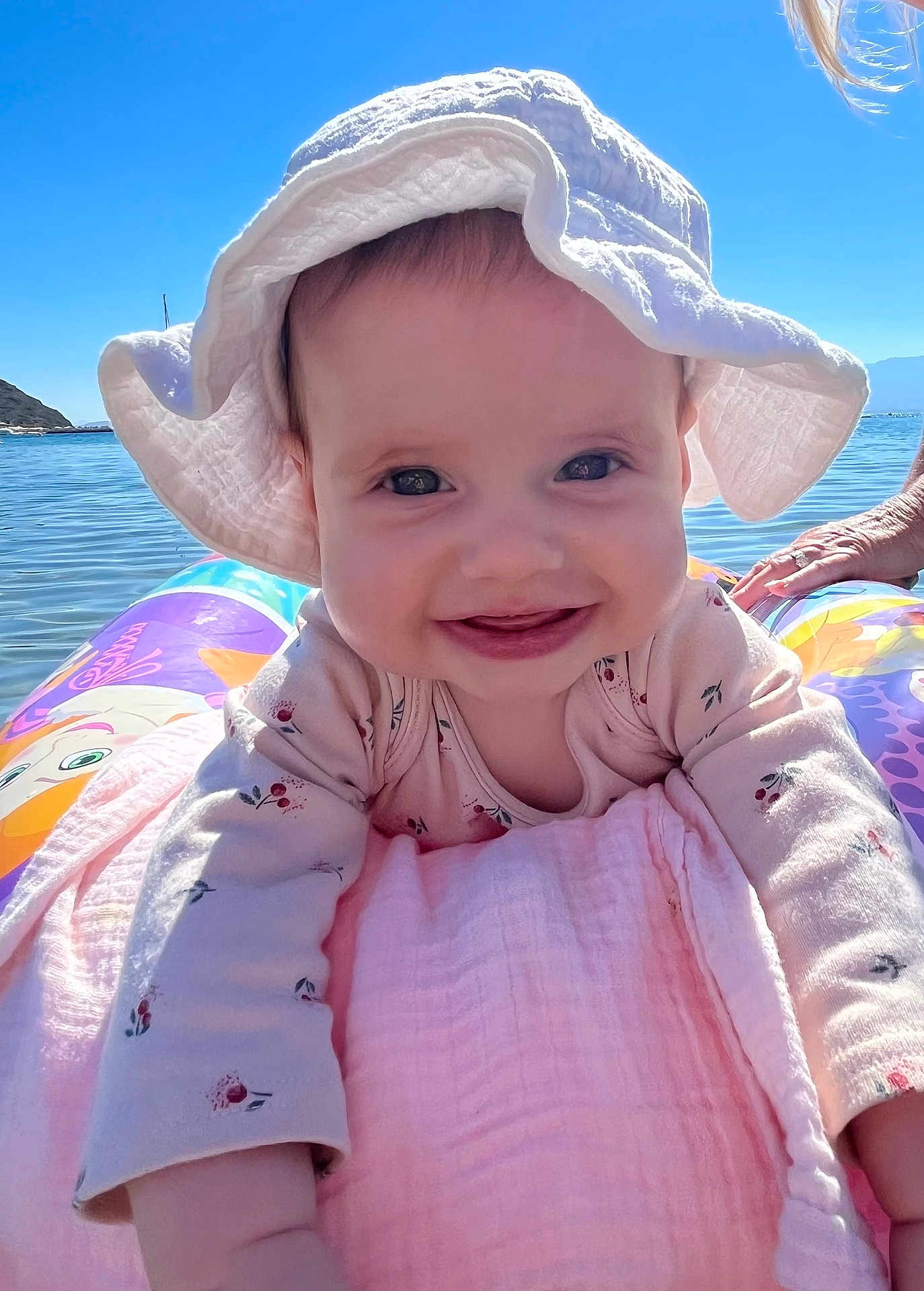 Isabelle joined the competition — help win amazing prizes! baby, child, smile, hat, sunhat, inflatable, water, lake, outdoor, sky, blue_sky, happy, face, person, summer, clothing, pink_blanket, nature, reaching, closeup