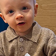 Diego participe au concours pour gagner de l'argent avec cette photo : blonde_hair, blue_eyes, brown_sweater, child, close_up, cozy_clothing, cup, curious, eyewear_absent, face, holding, indoor, person, portrait, seated, sweater, table, toddler, wooden_background, young_child