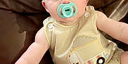 Zayden is registered to the contest to win money with this photo: baby, pacifier, romper, truck_design, couch, leather, child, infant, sitting, cute, soft_lighting, indoor, young, face, footwear, white_shoes, buttoned_clothing, plush, skin, portrait