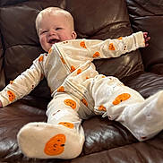Zayden is registered to the contest to win money with this photo: baby, child, smiling, pajamas, pumpkin_pattern, leather_couch, indoor, cute, happy, infant, person, clothing, footwear, relaxing, young_child, cozy, home, comfort, small_child, joyful