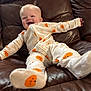 baby, child, smiling, pajamas, pumpkin_pattern, leather_couch, indoor, cute, happy, infant, person, clothing, footwear, relaxing, young_child, cozy, home, comfort, small_child, joyful