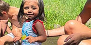 Nallely is registered to the contest to win money with this photo: child, girl, water, stream, outdoor, summer, nature, grass, rocks, playing, happy, smiling, pigtails, shorts, tank_top, sunlight, people, adult, boy, fun
