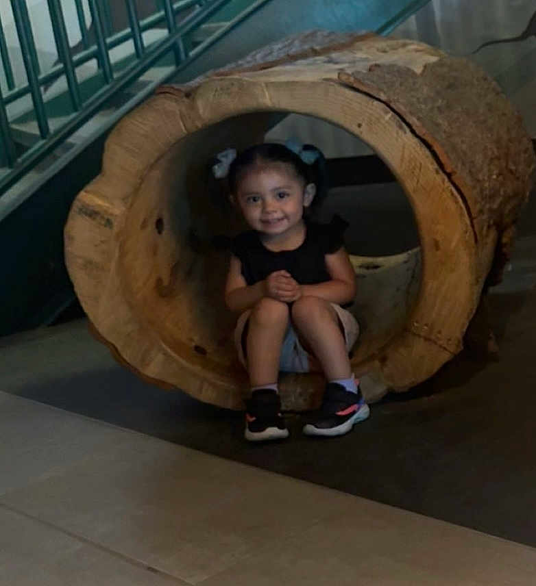 Nallely is registered to the contest to win money with this photo: child, girl, smile, pigtails, log, wood, indoor, floor, staircase, shoes, black_shirt, shorts, sitting, face, person, young, cute, happy, play, natural_material