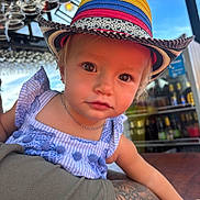 Olivia joined the competition — help win amazing prizes! toddler, child, hat, colorful_hat, blue_dress, tattoo, arm, glasses, outdoor, portrait, person, face, eyes, curious, table, sunlight, daylight, casual, restaurant, baby