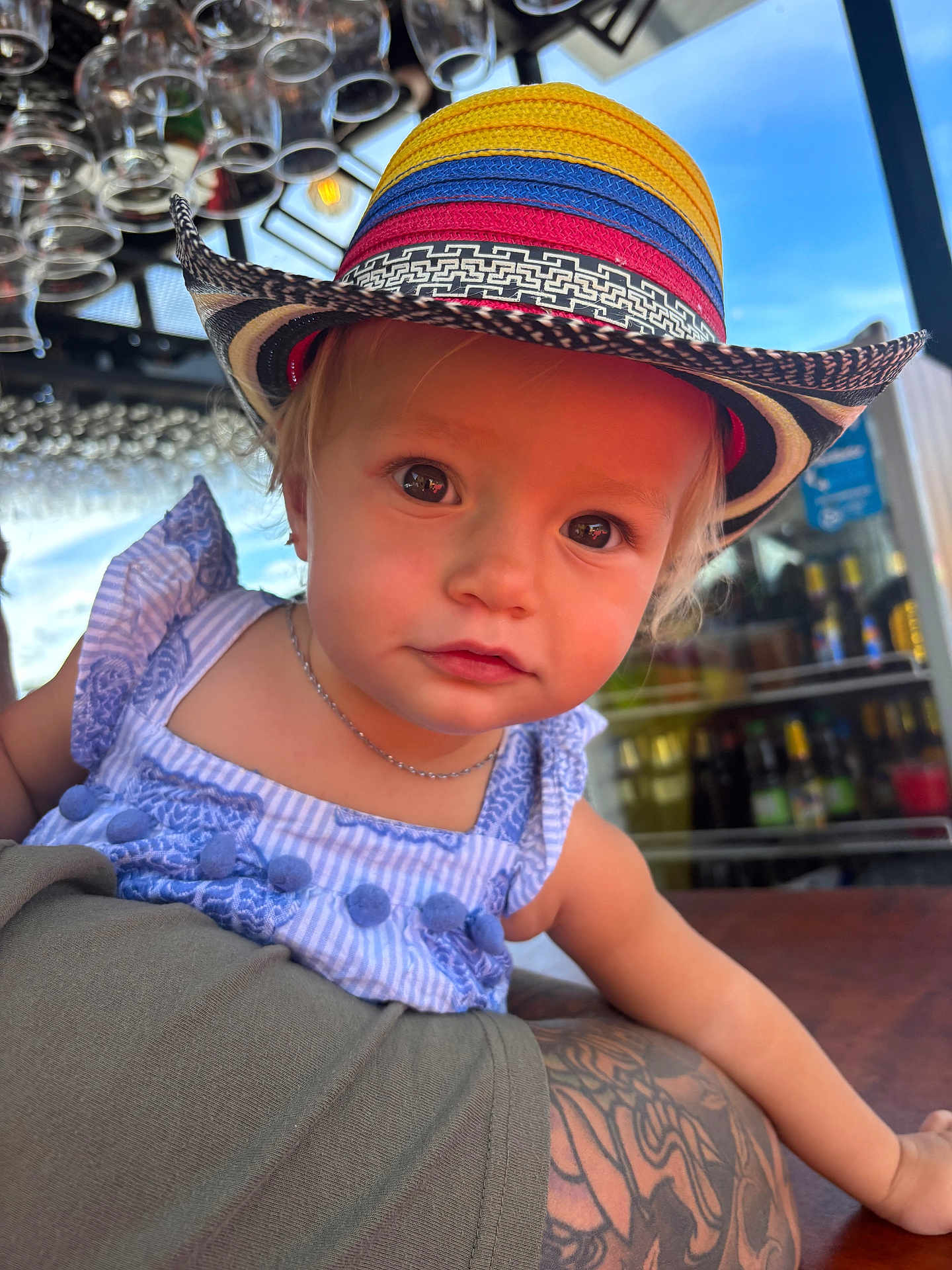 Olivia joined the competition — help win amazing prizes! toddler, child, hat, colorful_hat, blue_dress, tattoo, arm, glasses, outdoor, portrait, person, face, eyes, curious, table, sunlight, daylight, casual, restaurant, baby