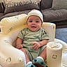 baby, baby_seat, bear_prints, child, clothing, comfortable, cozy, cushion, cute, dinosaur_slippers, happy, hat, indoor, infant, living_room, person, portrait, seated, smiling, soft_furniture