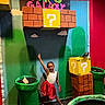 Tina is registered to the contest to win money with this photo: background, child, colorful, costume, decor, fun, girl, green_pipe, happy, indoor, nostalgia, playful, pose, question_mark_block, red_skirt, smile, standing, super_mario, video_game_theme, white_top