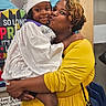 Tiana is registered to the contest to win money with this photo: celebration, child, classroom, colorful_background, education, event, family, graduation_cap, graduation_gown, happy, hug, indoor, kiss, person, portrait, proud, sign, smile, woman, yellow_dress