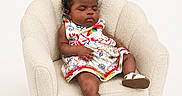 Tiana is registered to the contest to win money with this photo: baby, child, sleeping, armchair, dress, colorful_clothing, footwear, sandal, curly_hair, hair_accessory, indoor, furniture, cozy, minimalist, portrait, infant, cute, relaxed, peaceful, studio