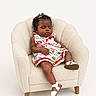 baby, child, sleeping, armchair, dress, colorful_clothing, footwear, sandal, curly_hair, hair_accessory, indoor, furniture, cozy, minimalist, portrait, infant, cute, relaxed, peaceful, studio