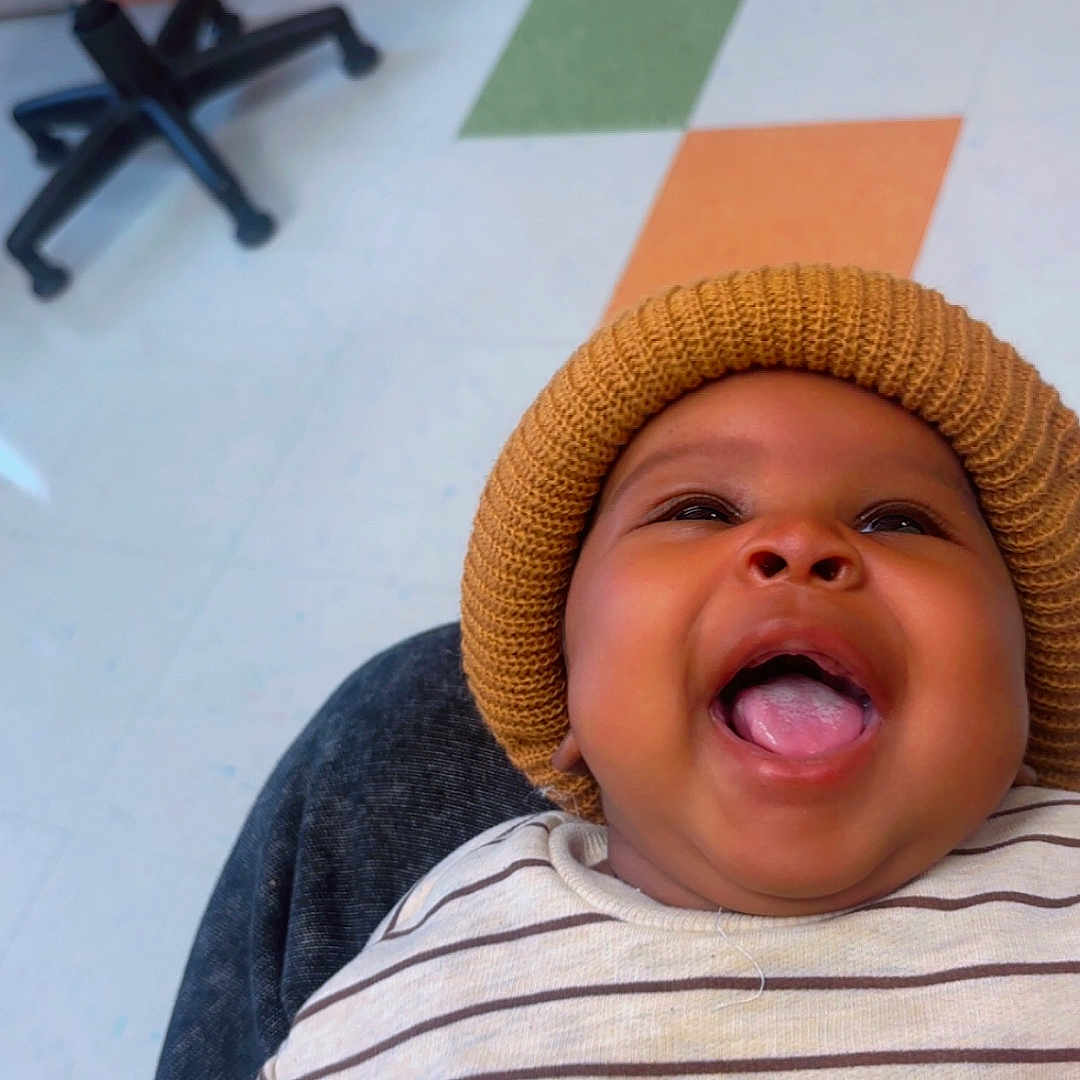 Tayvion Davis joined the competition — help win amazing prizes! baby, child, infant, smiling, laughing, portrait, close_up, knit_hat, striped_shirt, dog_design, clothing, sitting, indoor, office, chair, floor, colorful_tiles, home, happy, face