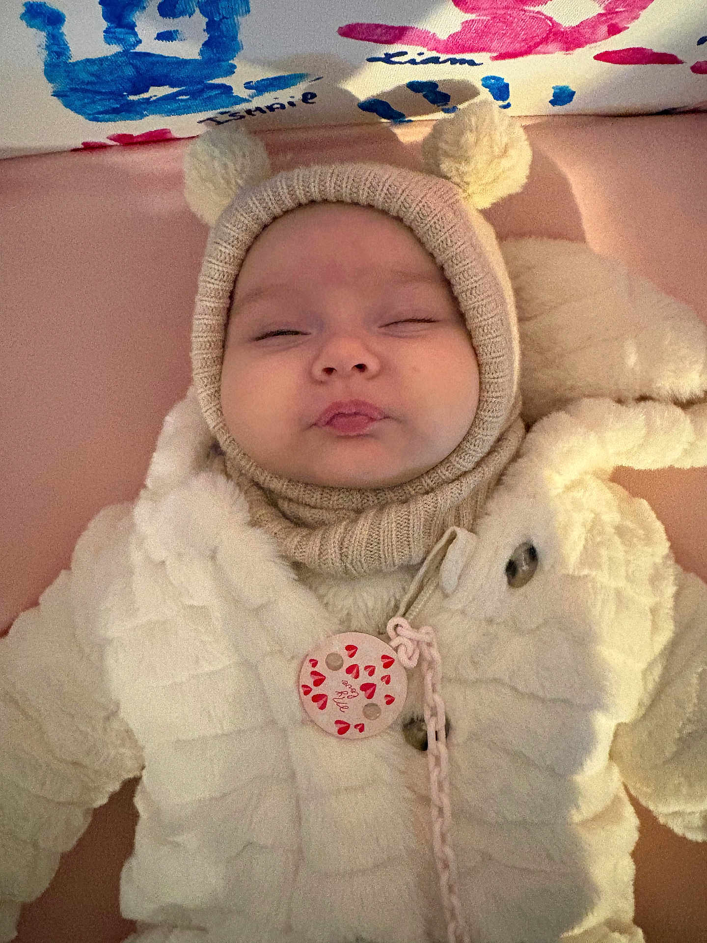 Arya participe au concours pour gagner de l'argent avec cette photo : baby, infant, child, coat, hat, pom_poms, knitwear, sleepy, face, pink_background, handprints, soft_texture, button, chain, warm_clothing, portrait, cute, indoors, smiling, person