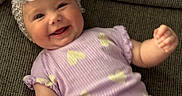 Charlotte is registered to the contest to win money with this photo: apparel, baby, child, couch, cozy, cute, flower, happy, headband, headwear, heart_pattern, indoor, infant, person, portrait, purple_clothing, relaxed, small_hands, smiling, texture