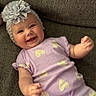 baby, smiling, headband, flower, purple_clothing, heart_pattern, couch, texture, infant, happy, portrait, child, cute, indoor, person, small_hands, cozy, relaxed, apparel, headwear