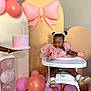 child, toddler, high_chair, birthday_cake, balloons, pink_dress, party, celebration, smiling, happy, indoor, decorations, person, footwear, hair_bows, table, pastel_colors, celebrant, event, presenting