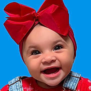 Charli is registered to the contest to win money with this photo: baby, child, infant, portrait, smiling, happy, red_bow, headband, blue_background, close_up, face, eyes, open_mouth, denim, overalls, red_clothing, floral_pattern, studio_photo, fashion, adorable