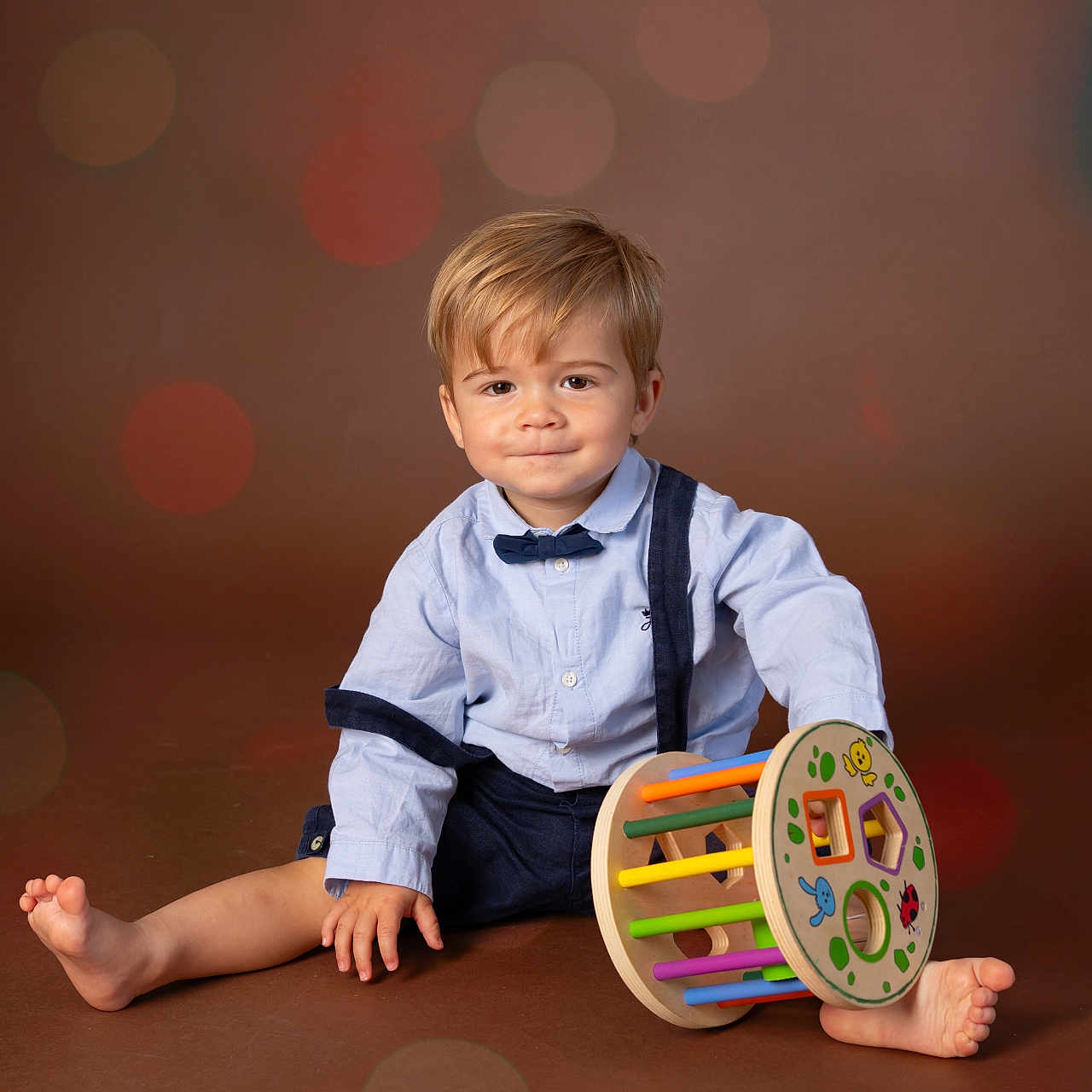 Jules Largeas participe au concours pour gagner de l'argent avec cette photo : barefoot, blue_shirt, bokeh, bow_tie, boy, brown_background, child, cute, floor, happy, indoor, play, portrait, sitting, smile, studio, suspenders, toddler, toy, wooden_toy