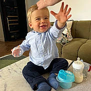 Jules Largeas participe au concours pour gagner de l'argent avec cette photo : toddler, child, baby, smiling, reaching_hand, adult_hand, blue_shirt, dark_pants, socks, baby_bottle, plastic_container, table, marble_surface, living_room, couch, pillow, floor, wooden_floor, indoor, happy