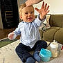 toddler, child, baby, smiling, reaching_hand, adult_hand, blue_shirt, dark_pants, socks, baby_bottle, plastic_container, table, marble_surface, living_room, couch, pillow, floor, wooden_floor, indoor, happy