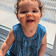 Bella is registered to the contest to win money with this photo: child, toddler, smiling, curly_hair, denim_dress, baby, portrait, indoor, sunlight, window, happy, face, cute, person, young_child, sitting, clothing, skin, hand, button