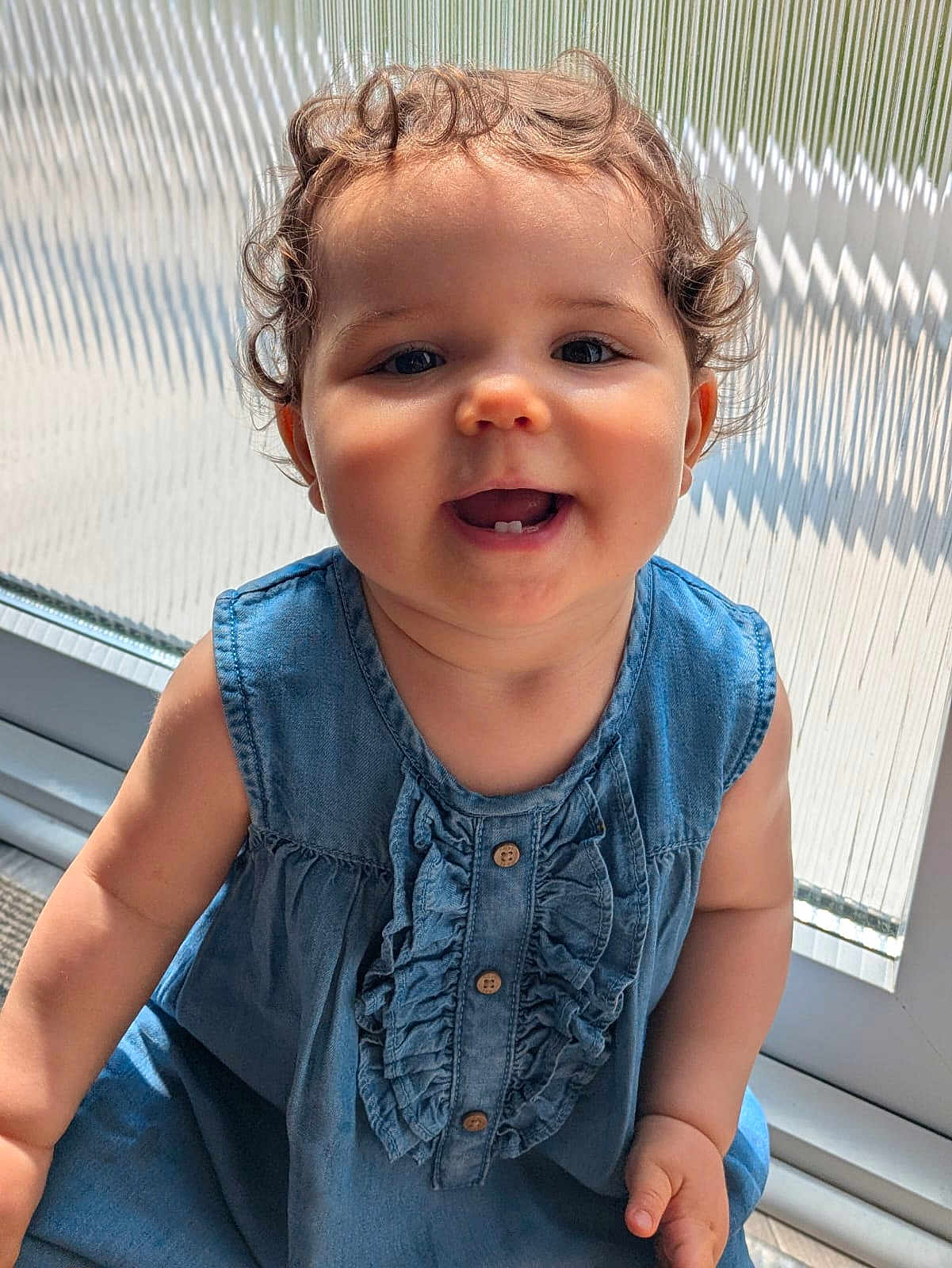 Bella is registered to the contest to win money with this photo: child, toddler, smiling, curly_hair, denim_dress, baby, portrait, indoor, sunlight, window, happy, face, cute, person, young_child, sitting, clothing, skin, hand, button
