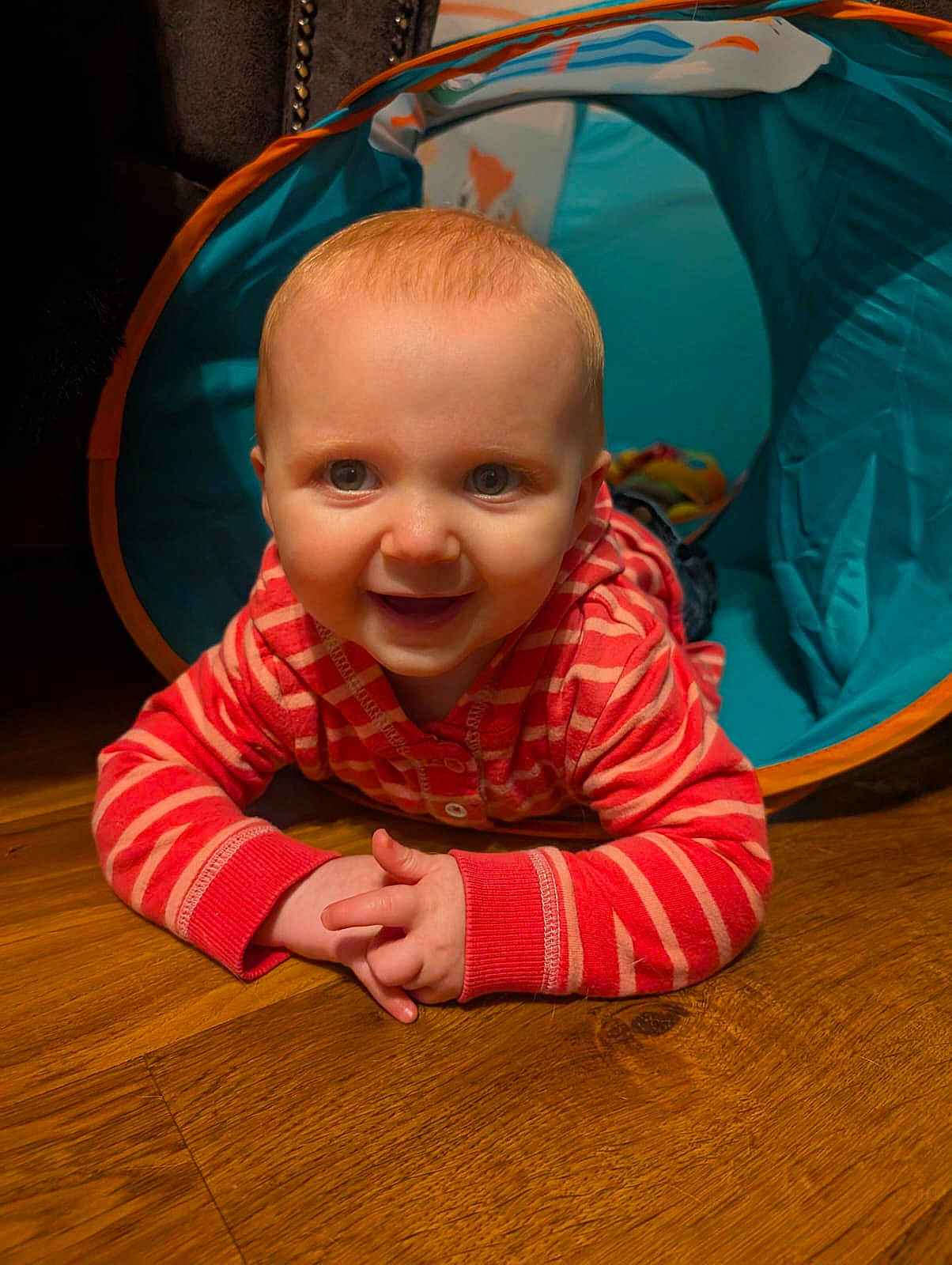Evie is registered to the contest to win money with this photo: baby, child, smiling, crawling, play_tunnel, striped_clothing, hoodie, wooden_floor, indoor, happy, cute, person, face, blue, red, young_child, infant, hands, eyes, play