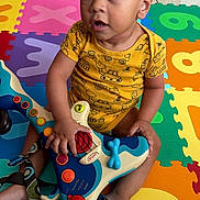 Ravi is registered to the contest to win money with this photo: baby, child, infant, portrait, sitting, wide_eyed, curly_hair, smiling, musical_toy, toy, play_mat, alphabet_mat, colorful, indoors, floor, onesie, barefoot, playtime, home, safe