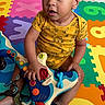 baby, child, infant, portrait, sitting, wide_eyed, curly_hair, smiling, musical_toy, toy, play_mat, alphabet_mat, colorful, indoors, floor, onesie, barefoot, playtime, home, safe