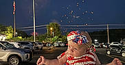 Marilyn is registered to the contest to win money with this photo: baby, child, patriotic, dress, headband, fireworks, night, parking_lot, car, american_flag, person, celebration, outdoor, holiday, cute, festive, holding_hands, sky, lights, people