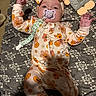 baby, bed, blanket, child, cozy, floral_pattern, flowers, halloween, headband, indoors, infant, lying_down, onesie, pacifier, plush_toy, portrait, pumpkin, smiling, text_on_toy, toy