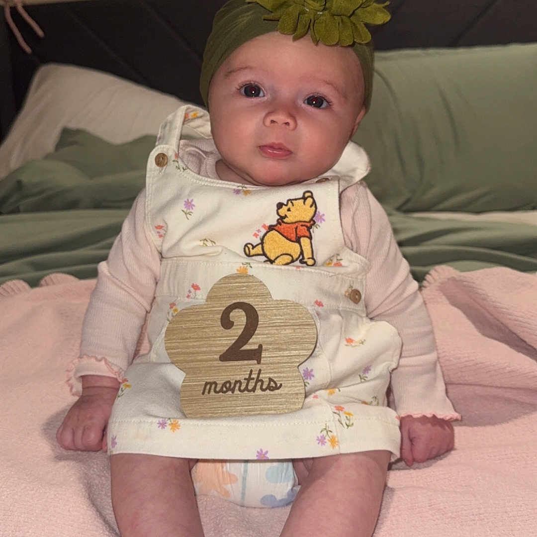 Marilyn joined the competition — help win amazing prizes! baby, infant, child, sitting, blanket, headband, flower, dress, winnie_the_pooh, wooden_sign, bed, pillow, diaper, indoor, cute, portrait, soft_lighting, pink, green, two_months