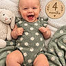 baby, blanket, carpet, child, close_up, daisy_pattern, family_photo, floral_clothing, four_months, green_outfit, happy, indoor, infant, lying_down, milestone_sign, portrait, rabbit, smiling, stuffed_animal, wooden_sign