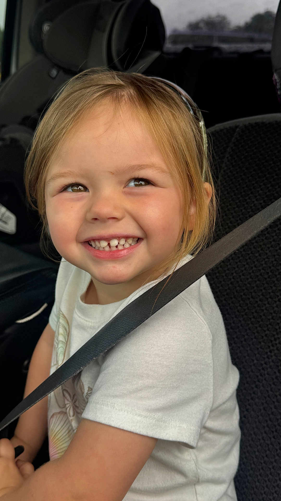 Reese is registered to the contest to win money with this photo: child, smiling, seatbelt, car_seat, vehicle, happy, young, girl, casual_clothing, short_sleeves, light_hair, indoors, portrait, closeup, safety, seat, car_interior, cute, teeth, joy