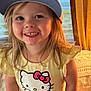 child, smiling, hat, baseball_cap, hello_kitty, shirt, yellow_shirt, pink_shorts, indoor, carpet, curtain, person, happy, face, long_hair, standing, cute, childhood, apparel, portrait