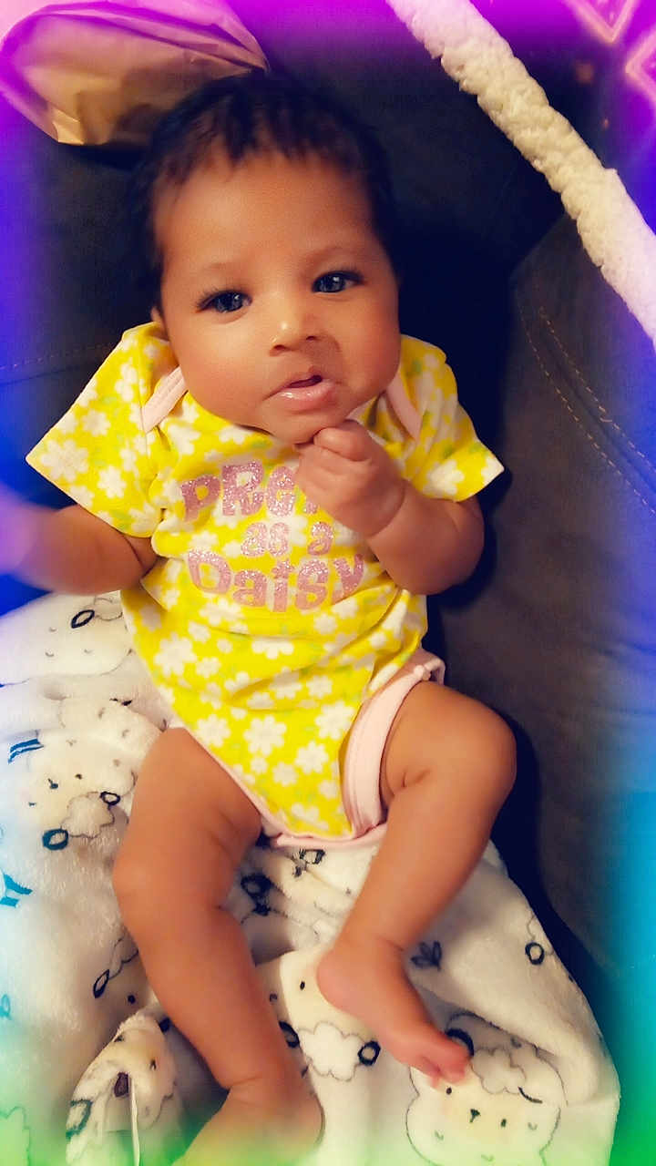 Lil Gbaby joined the competition — help win amazing prizes! baby, infant, yellow_onesie, blanket, sheep_pattern, cute, child, portrait, indoor, soft_lighting, hand, face, legs, cozy, comfortable, expression, colorful_vignette, relaxed, young_child, seated