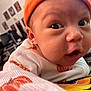 baby, infant, close_up, face, orange_hat, white_sweater, curious_expression, indoor, blurred_background, furniture, wall_art, child, person, portrait, cute, wide_eyes, open_mouth, cozy, warm_colors, knitted_clothing