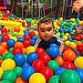 ball_pit, blue_balls, blue_shirt, bright_colors, casual_clothing, child, colorful_balls, curly_hair, fun, green_balls, hands, indoor_playground, orange_balls, play_area, playtime, red_balls, smiling, toddler, toy, yellow_balls
