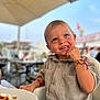 blue_eyes, blurred_background, bracelet, cafe, casual_clothing, child, daylight, eating, food, french_fries, happy, high_chair, outdoor, person, plate, shirt, smiling, table, toddler, umbrella