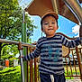 blue_sky, boy, cap, casual_clothing, child, clouds, daylight, fence, grass, greenery, happy, metal_bars, outdoor, person, play_equipment, playground, smiling, striped_shirt, trees, vehicle
