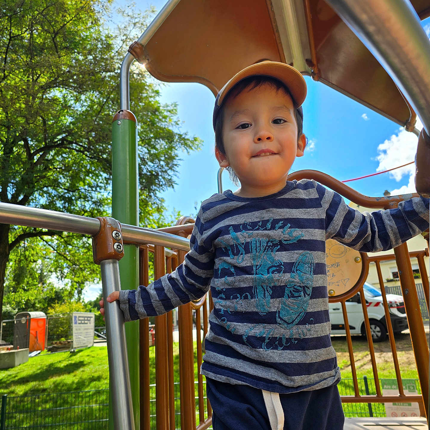 Devy Hong Yi participe au concours pour gagner de l'argent avec cette photo : blue_sky, boy, cap, casual_clothing, child, clouds, daylight, fence, grass, greenery, happy, metal_bars, outdoor, person, play_equipment, playground, smiling, striped_shirt, trees, vehicle