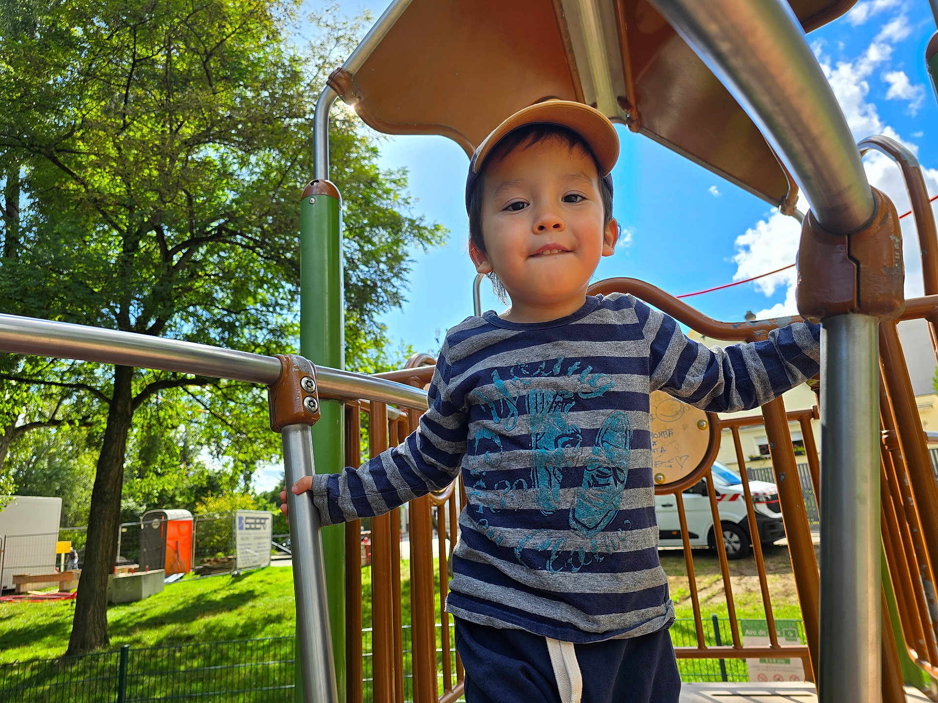 Devy Hong Yi participe au concours pour gagner de l'argent avec cette photo : blue_sky, boy, cap, casual_clothing, child, clouds, daylight, fence, grass, greenery, happy, metal_bars, outdoor, person, play_equipment, playground, smiling, striped_shirt, trees, vehicle