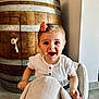 toddler, child, smiling, happy, plush_toy, elephant_rocker, bow, hair_accessory, white_shirt, wooden_barrel, indoor, floor, tile_floor, furniture, cute, person, baby, playful, joyful, seat