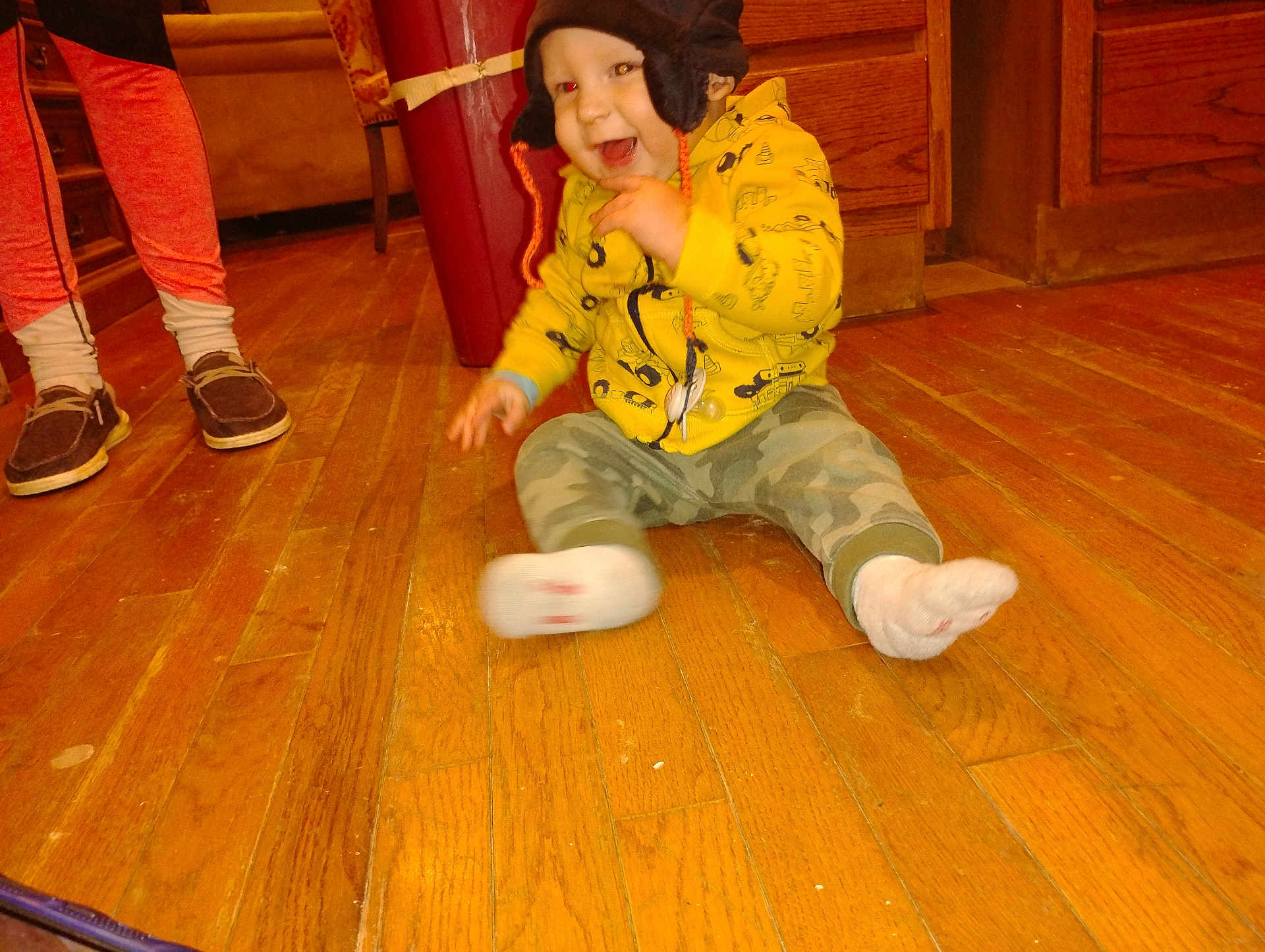 Steve is registered to the contest to win money with this photo: toddler, child, yellow_hoodie, hat, floor, wooden_floor, camo_pants, socks, smile, indoor, person, legs, shoes, moccasin, happy, motion_blur, wood_cabinet, furniture, warm_clothing, playful
