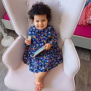 Inès a rejoint le concours — aidez-le/la à gagner de superbes lots ! child, toddler, smiling, curly_hair, floral_dress, armchair, pink_armchair, wafer_biscuit, smartphone, indoors, wooden_floor, cozy_room, furniture, sitting, happy, cute, person, playful, relaxed, portrait