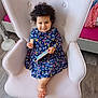 child, toddler, smiling, curly_hair, floral_dress, armchair, pink_armchair, wafer_biscuit, smartphone, indoors, wooden_floor, cozy_room, furniture, sitting, happy, cute, person, playful, relaxed, portrait