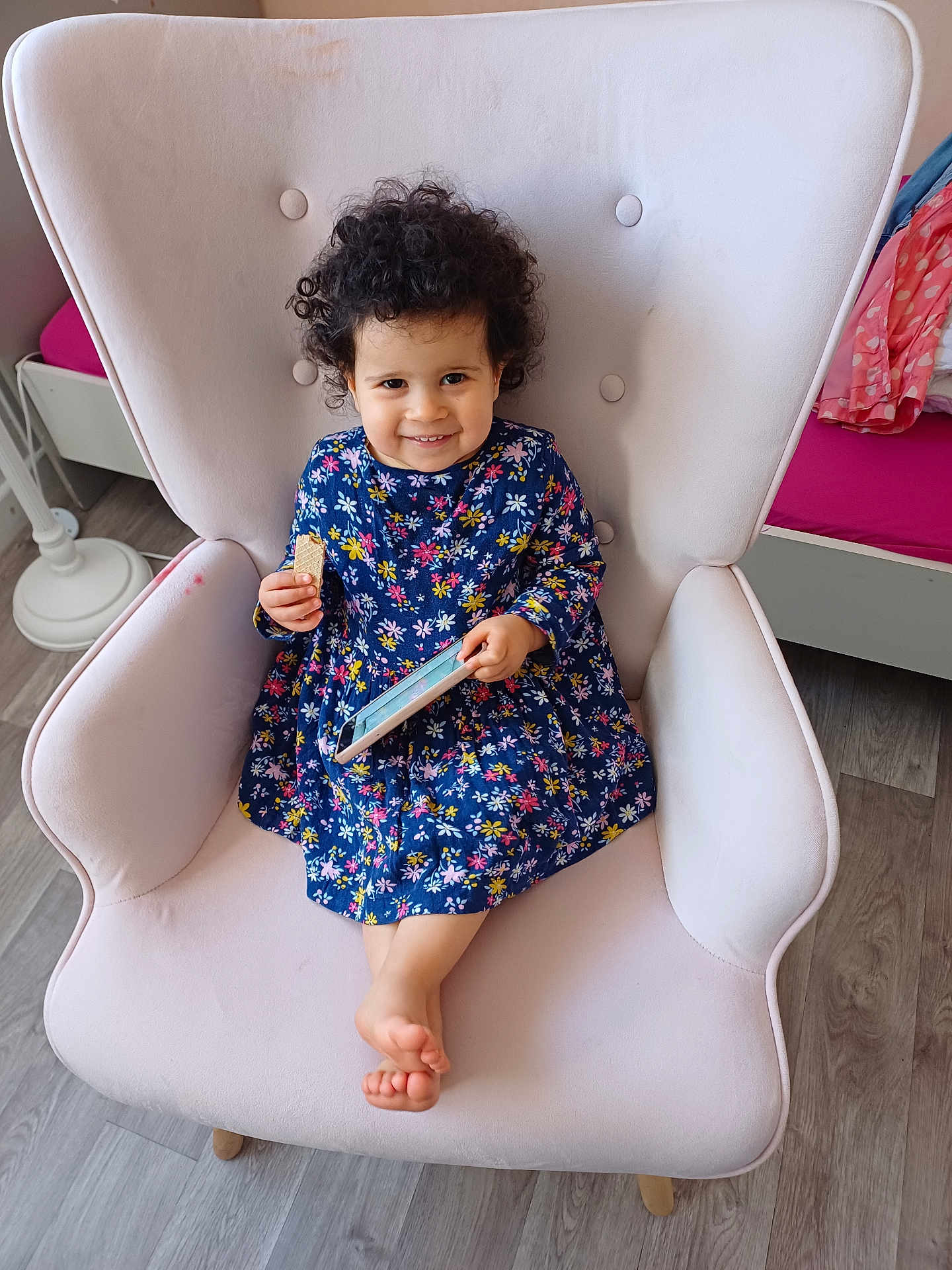 Inès a rejoint le concours — aidez-le/la à gagner de superbes lots ! child, toddler, smiling, curly_hair, floral_dress, armchair, pink_armchair, wafer_biscuit, smartphone, indoors, wooden_floor, cozy_room, furniture, sitting, happy, cute, person, playful, relaxed, portrait