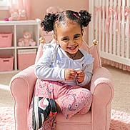 Ilaria is registered to the contest to win money with this photo: child, girl, smiling, pigtails, pink, armchair, nursery, crib, plush_toy, carpet, sneakers, clothing, room, wall_art, curtains, furniture, happy, indoor, person, portrait