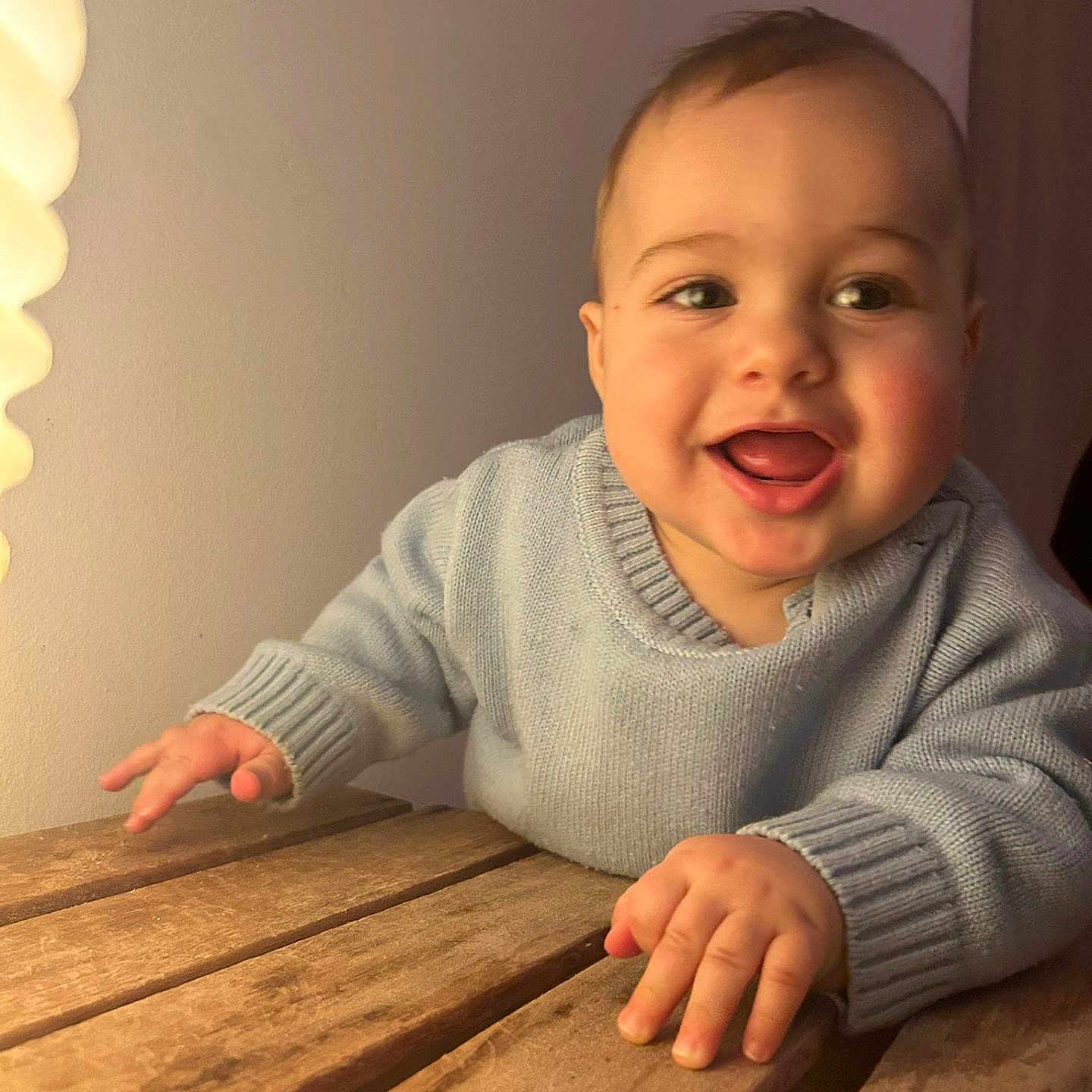 Loukas a rejoint le concours — aidez-le/la à gagner de superbes lots ! baby, blue_sweater, cheeks, close_up, crawling, eyes, face, furniture, hands, happy, indoor, infant, lamp, mouth, portrait, skin, smile, table, warm_light, wooden_table