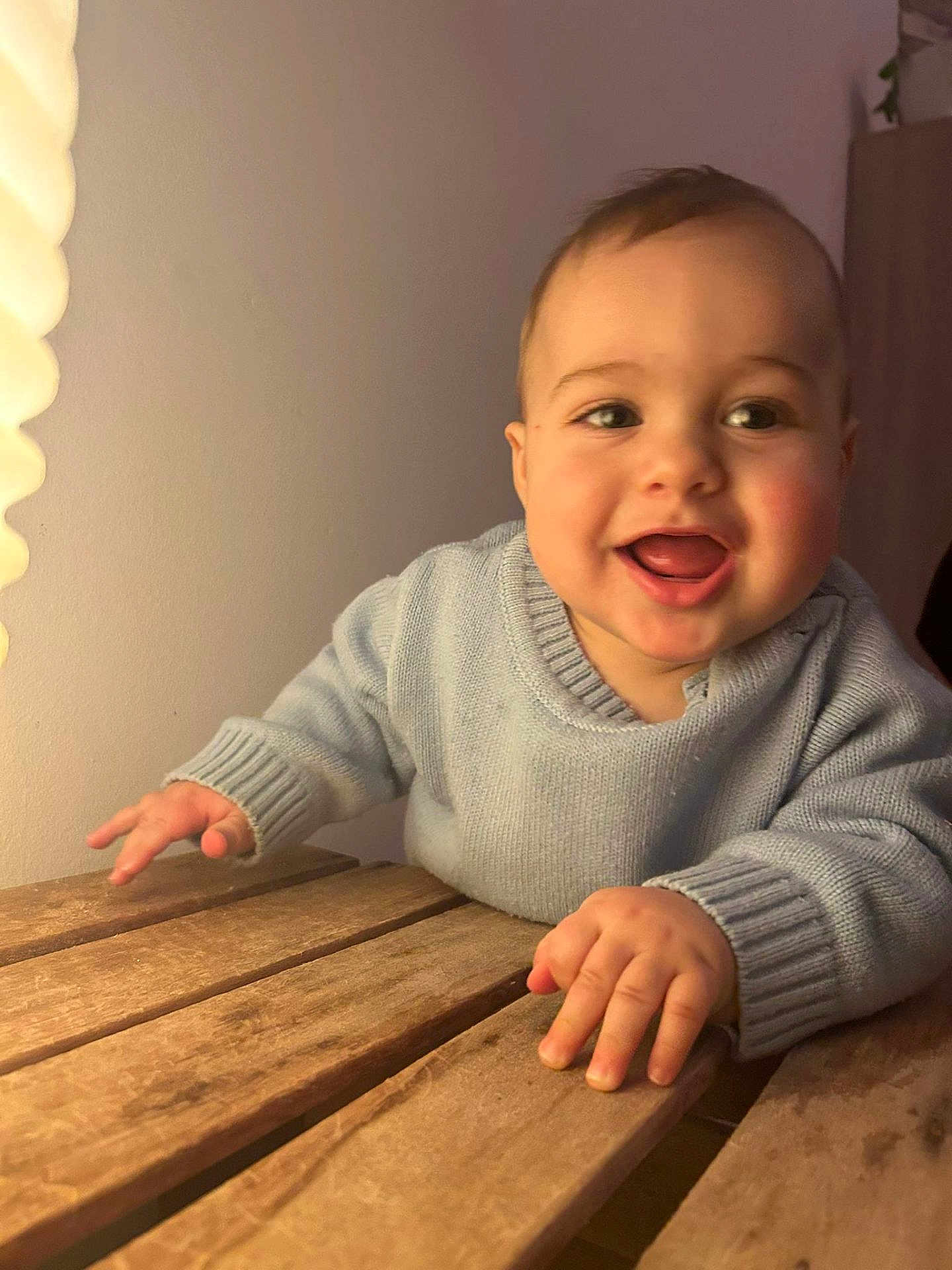 Loukas a rejoint le concours — aidez-le/la à gagner de superbes lots ! baby, infant, smile, happy, blue_sweater, hands, wooden_table, table, indoor, warm_light, cheeks, eyes, mouth, face, portrait, close_up, crawling, furniture, lamp, skin